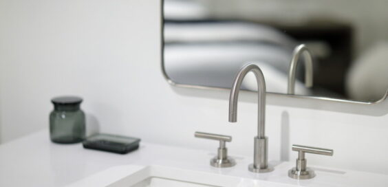 A stainless steel faucet and sink. There is a mirror above the sink and decorative objects on the white counter.