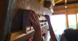 A close-up view of a woman installing a fingerboard trainer onto a gray indoor rock climbing wall in her home.