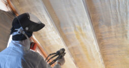 A person wearing personal protective equipment using a spray foam machine to apply insulation to a ceiling.