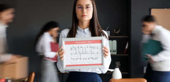 A woman wearing business casual attire holds a fire evacuation building plan while blurred people walk behind her.