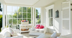 A bright, sunny conservatory with white walls and furniture and a large panel window facing tall green trees.