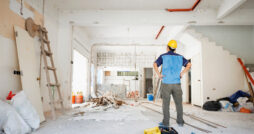 A man with a hard hat on looks at the interior of a home that is being renovated. Material and tools are against the walls.