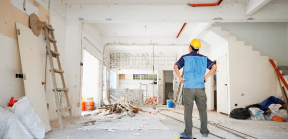 A man with a hard hat on looks at the interior of a home that is being renovated. Material and tools are against the walls.