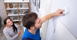 A female homeowner watches a repair man stand on a ladder and test the smoke detector he's just installed.