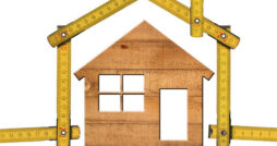 A small wooden model of a house standing against a white background and surrounded by an intricate folding ruler.