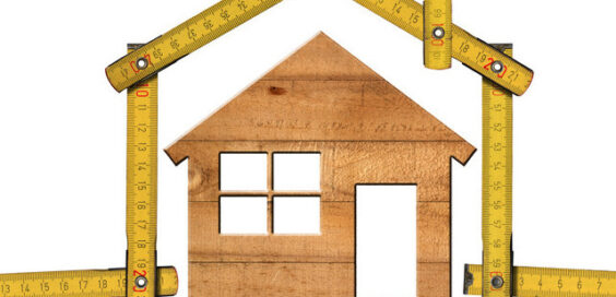 A small wooden model of a house standing against a white background and surrounded by an intricate folding ruler.