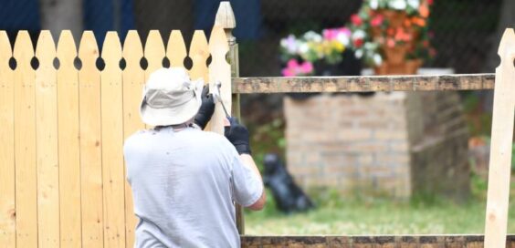 A worker wearing protective clothing for outdoor garden work. He is installing a wooden fence using nails and a hammer.
