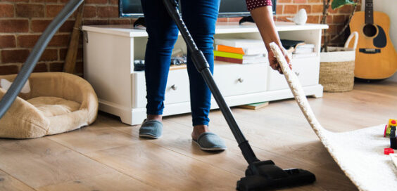 A woman wearing slippers lifts a corner of an area rug while she vacuums the hardwood floors in her living room.