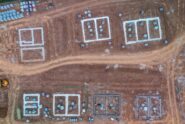 An aerial view of a flat dirt construction site featuring seven home foundations and construction materials lying around.