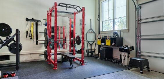 A gym setup with a black mat, free wieghts, a stepping stool, and still rings in a garage with windows.
