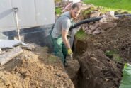 A young man wearing green overalls and yellow gloves uses a shovel to dig a large hole in front of a pipe.