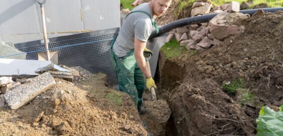 A young man wearing green overalls and yellow gloves uses a shovel to dig a large hole in front of a pipe.