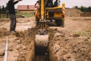 Construction worker standing next to a yellow crane truck digging a ditch in an empty lot covered in soil.