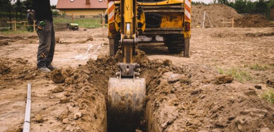 Construction worker standing next to a yellow crane truck digging a ditch in an empty lot covered in soil.