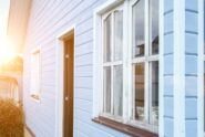 A close-up of a small dwelling unit with blue siding, white rustic windows, and an open brown front door.
