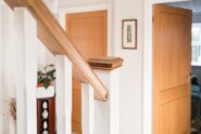A close-up of a wooden staircase handrail inside a home with white painted walls and warm wood accents.