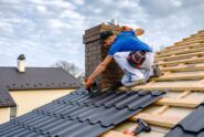 A construction worker wearing a blue shirt sits on the roof of a house while nailing down metal shingles.