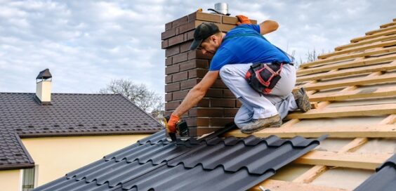 A construction worker wearing a blue shirt sits on the roof of a house while nailing down metal shingles.