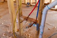Two PEX tubes and a PVC drain pipe are secured to lumber above wood shavings, with yellow wiring nearby.