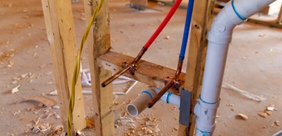 Two PEX tubes and a PVC drain pipe are secured to lumber above wood shavings, with yellow wiring nearby.