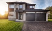 A modern home with a gray brick facade, brown siding, black garage doors, and a freshly paved driveway.