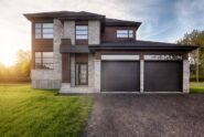A modern home with a gray brick facade, brown siding, black garage doors, and a freshly paved driveway.