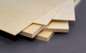 A close up of the corner of a stack of plywood. There are various styles and thicknesses of plywood on a gray surface.