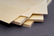 A close up of the corner of a stack of plywood. There are various styles and thicknesses of plywood on a gray surface.