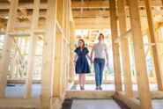 A young couple smiling as they walk through the wooden studs and frame of a new home under construction.