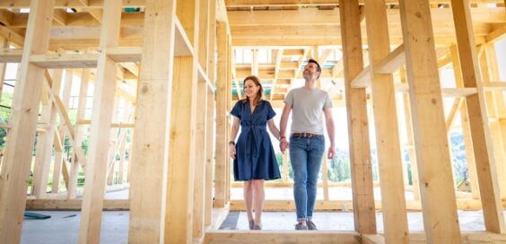 A young couple smiling as they walk through the wooden studs and frame of a new home under construction.