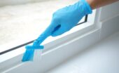 A person wearing blue latex gloves cleans the frame of a sliding glass door with a brush, leaving the area spotless.