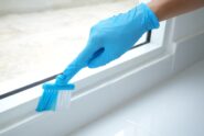 A person wearing blue latex gloves cleans the frame of a sliding glass door with a brush, leaving the area spotless.