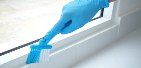 A person wearing blue latex gloves cleans the frame of a sliding glass door with a brush, leaving the area spotless.