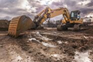An excavator shoveling wet dirt and mud at a construction site. The sky is full of scattered, dark rainclouds.