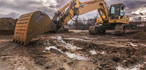 An excavator shoveling wet dirt and mud at a construction site. The sky is full of scattered, dark rainclouds.