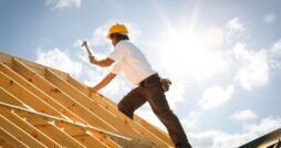 A man wearing a white shirt and a hard hat hammers through the wooden frame of a home's roof.