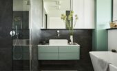 A small minimalist bathroom features a white tub, black shower surround, green walls, cabinetry, and accents.