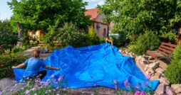 Two people spreading a large blue pond liner in a backyard surrounded by flowers, bushes, and trees on a sunny day.