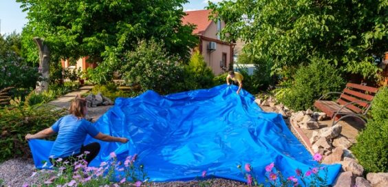 Two people spreading a large blue pond liner in a backyard surrounded by flowers, bushes, and trees on a sunny day.