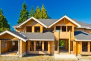 A house under construction stands with a finished gray roof. Behind the house are trees, and a clear blue sky above.