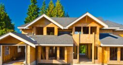 A house under construction stands with a finished gray roof. Behind the house are trees, and a clear blue sky above.