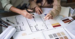 Two people sit at a desk covered with home building materials, including blueprints, paint swatches, and wood samples.