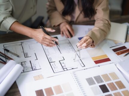 Two people sit at a desk covered with home building materials, including blueprints, paint swatches, and wood samples.