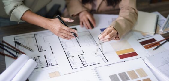 Two people sit at a desk covered with home building materials, including blueprints, paint swatches, and wood samples.