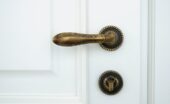 A white wooden door with an ornate handle doorknob. A lock mechanism is beneath it. Both fixtures are made of bronze.