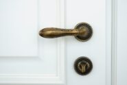 A white wooden door with an ornate handle doorknob. A lock mechanism is beneath it. Both fixtures are made of bronze.