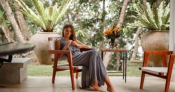 A woman wearing a long gray dress sits on a wooden chair next to a table with a glass flower vase on top of it.