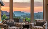 Two accent chairs sit in front of floor-to-ceiling picture windows with a view of a sunset over a mountain range.