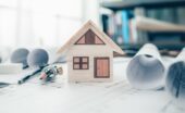 A small, wooden model house is sitting on top of a paper, alongside rolled up blueprints and drawing tools.
