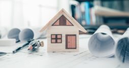 A small, wooden model house is sitting on top of a paper, alongside rolled up blueprints and drawing tools.